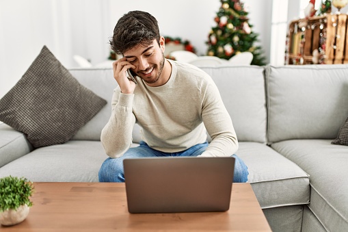 Young hispanic man sitting on the sofa using laptop and smartphone at home.