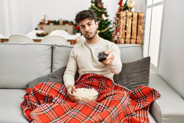 istock Young hispanic man eating popcorn sitting on sofa by christmas tree thinking attitude and sober expression looking self confident 1346561488