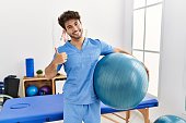 Hispanic physiotherapy man working at pain recovery clinic smiling happy and positive, thumb up doing excellent and approval sign
