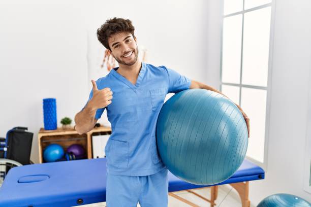 Hispanic physiotherapy man working at pain recovery clinic smiling happy and positive, thumb up doing excellent and approval sign stock photo