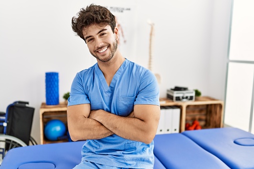 Young hispanic man wearing physio therapist uniform with arms crossed gesture at clinic