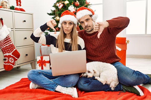 Young couple of wife and husband wearing christmas hat using laptop with angry face, negative sign showing dislike with thumbs down, rejection concept