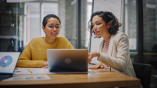 Two Diverse Multiethnic Female Have a Discussion in Meeting Room Behind Glass Walls in an Agency. Creative Director and Project Manager Compare Business Results on Laptop and App Designs in an Office.