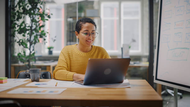 retrato de jovem especialista em marketing latino em óculos trabalhando em computador portátil em ambiente ocupado de escritório criativo. beautiful diverse multiethnic female manager está navegando na internet. - adult adults only assistant occupation - fotografias e filmes do acervo