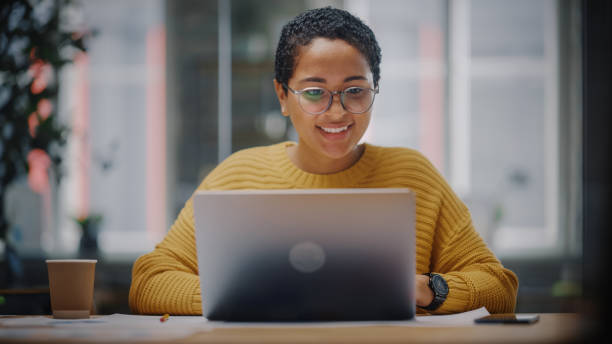 feliz especialista en marketing latino en gafas trabajando en computadora portátil en oficina creativa ocupada. hermosa y diversa gerente de proyecto dudando y pensando antes de responder un correo electrónico a un colega. - hesitating fotografías e imágenes de stock