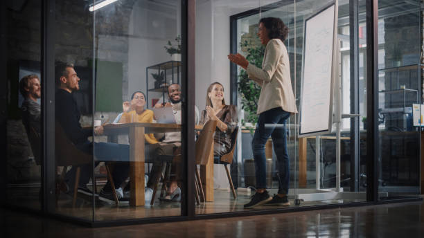 istock Project Manager Makes a Presentation for a Young Diverse Creative Team in Meeting Room in an Agency. Colleagues Sit Behind Conference Table and Discuss Business Development, User Interface and Design. 1346563249