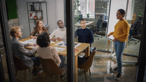 istock Project Manager Makes a Presentation for a Young Diverse Creative Team in Meeting Room in an Agency. Colleagues Sit Behind Conference Table and Discuss Business Development, User Interface and Design. 1346563263