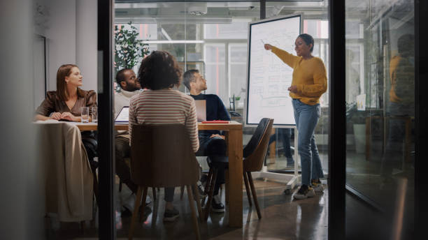 istock Project Manager Makes a Presentation for a Young Diverse Creative Team in Meeting Room in an Agency. Colleagues Sit Behind Conference Table and Discuss Business Development, User Interface and Design. 1346563269