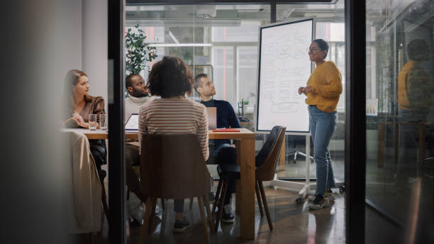 istock Project Manager Makes a Presentation for a Young Diverse Creative Team in Meeting Room in an Agency. Colleagues Sit Behind Conference Table and Discuss Business Development, User Interface and Design. 1346563273
