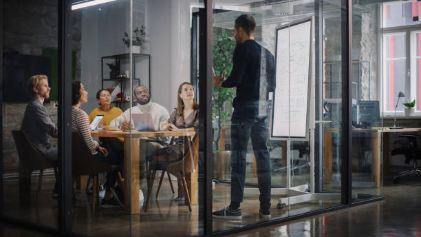 istock Project Manager Makes a Presentation for a Young Diverse Creative Team in Meeting Room in an Agency. Colleagues Sit Behind Conference Table and Discuss Business Development, User Interface and Design. 1346563292