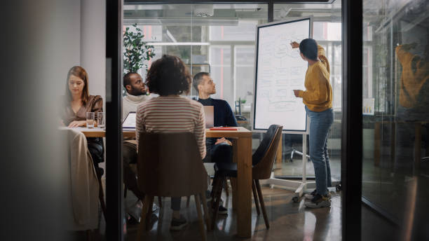 istock Project Manager Makes a Presentation for a Young Diverse Creative Team in Meeting Room in an Agency. Colleagues Sit Behind Conference Table and Discuss Business Development, User Interface and Design. 1346563293