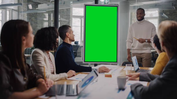 istock Project Manager Makes a Presentation on a Green Screen Mock Up Display for a Young Diverse Creative Team in Meeting Room in an Agency. Colleagues Sit Behind Conference Table and Discuss Business. 1346563357