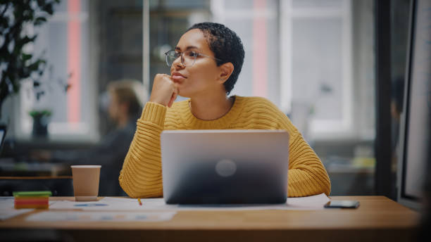 feliz especialista en marketing latino en gafas trabajando en computadora portátil en oficina creativa ocupada. hermosa y diversa gerente de proyecto dudando y pensando antes de responder un correo electrónico a un colega. - hesitating fotografías e imágenes de stock