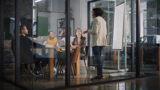istock Project Manager Makes a Presentation for a Young Diverse Creative Team in Meeting Room in an Agency. Colleagues Sit Behind Conference Table and Discuss Business Development, User Interface and Design. 1346563378