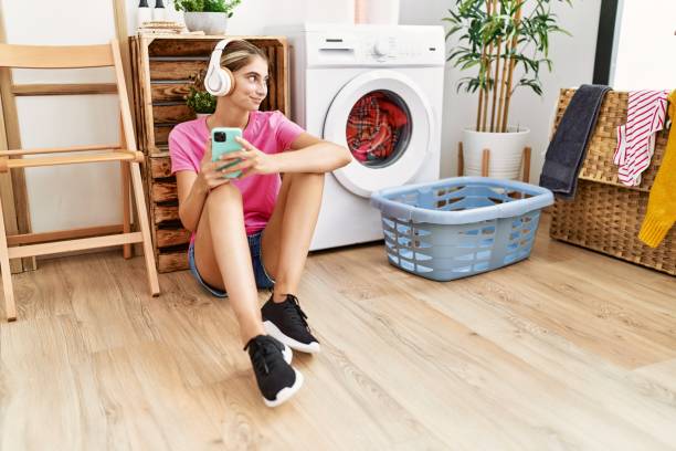 joven caucásica con teléfono inteligente y auriculares esperando la lavadora en la lavandería - all laundry detergent audio fotografías e imágenes de stock