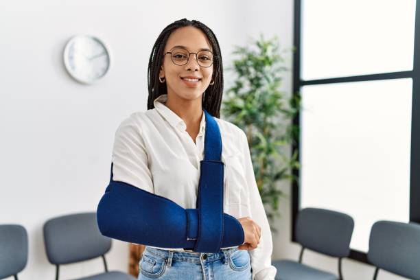 joven afroamericana sonriendo con confianza lesión en el brazo en la sala de espera de la clínica - arm sling fotografías e imágenes de stock