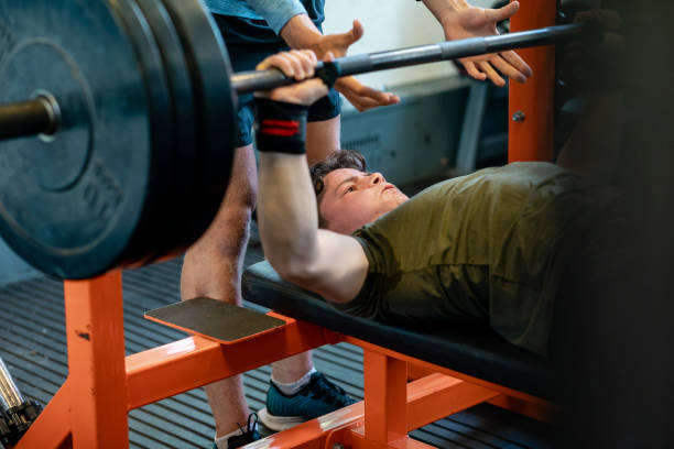 Striving to Achieve A close-up shot of a young teenage male athlete performing a bench press with a barbell, he is taking guidance from a fitness instructor, who is squatting him. He is lying down on a bench, retracting his scapular and using his pectoral muscles to lower and raise the weighted bar. LIFTING WEIGHTS stock pictures, royalty-free photos & images