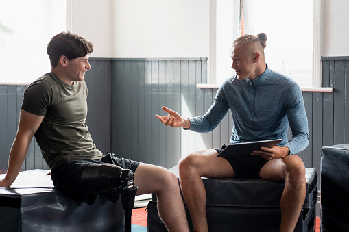 A front-view shot of a young teenage male athlete with a prosthetic leg taking guidance from a fitness instructor who is using a digital tablet in a gym.