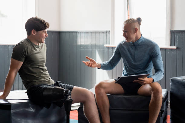 We're Going to Have a Good Session Today! A front-view shot of a young teenage male athlete with a prosthetic leg taking guidance from a fitness instructor who is using a digital tablet in a gym. fitness trainer stock pictures, royalty-free photos & images