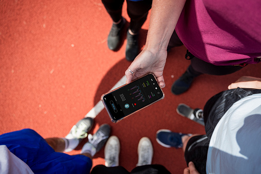 A close-up elevated view shot of a group of unrecognizable friends including a young teenage male athlete with a prosthetic leg standing on a running track, they are using a smartphone to check a fitness app after a cardio training session.
