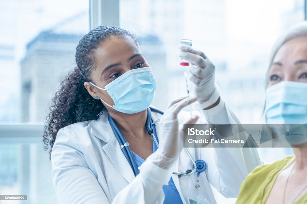 Doctor Filling a Syringe from a Vial A female doctor fills a syringe from a vaccination vial as her senior patient looks away anxiously.  Both are wearing medical masks to protect them from COVID. Vaccination Stock Photo