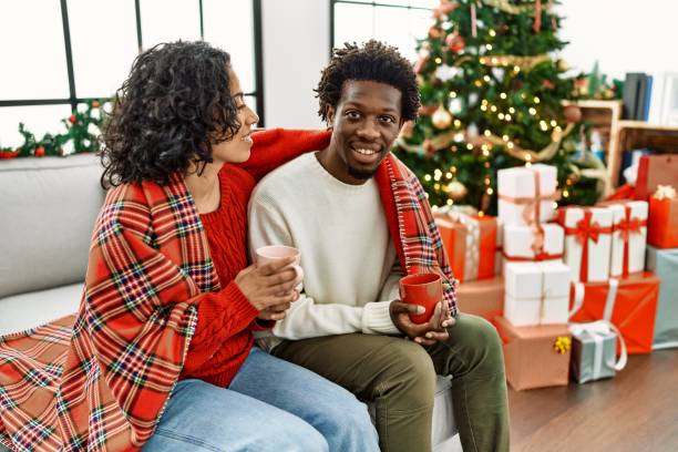istock Young interracial couple drinking coffee sitting on the sofa by christmas tree at home. 1346566741