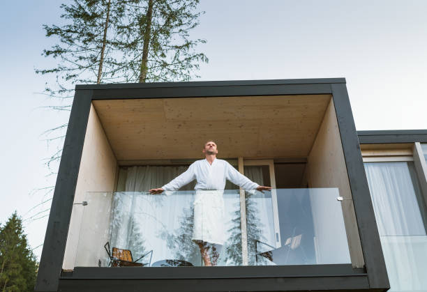 istock Resting Man dressed white bathrobe standing on forest house balcony and enjoying fresh air with nature forest pine trees view. Outside Scandinavian interior design house. Living in wild concept image 1346567469