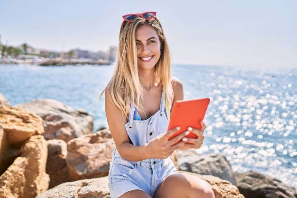 istock Young blonde girl using touchpad sitting on the rock at the beach. 1346567562