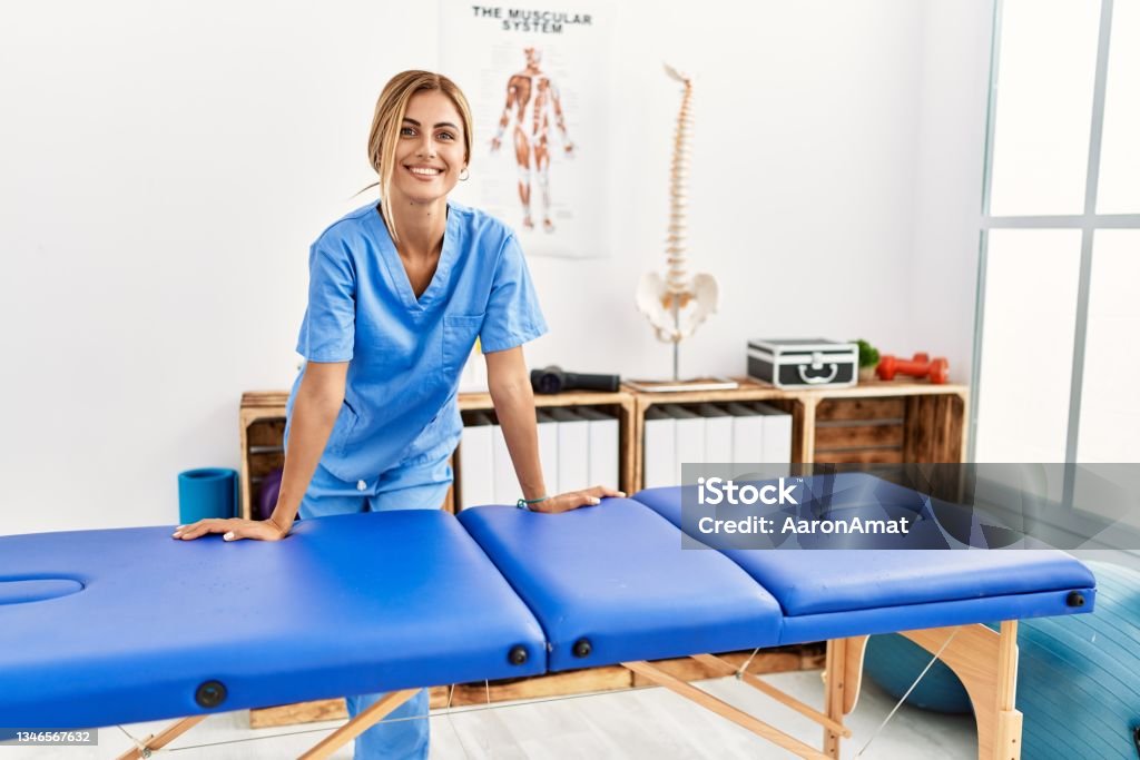 Young caucasian physio therapist smiling happy leaning on table at the clinic Massage Therapist Stock Photo