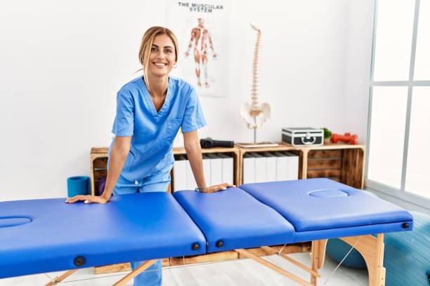 jovem fisioterapeuta caucasiano sorrindo feliz inclinando-se sobre a mesa na clínica - massage therapist - fotografias e filmes do acervo