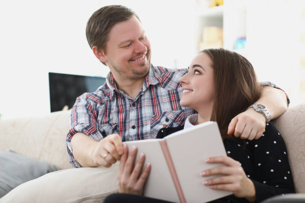 istock Young married couple sitting on sofa with book and looking at each other 1346568739