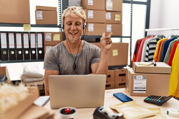 istock Young blond man wearing operator headset working at online shop smiling with happy face winking at the camera doing victory sign. number two. 1346568828
