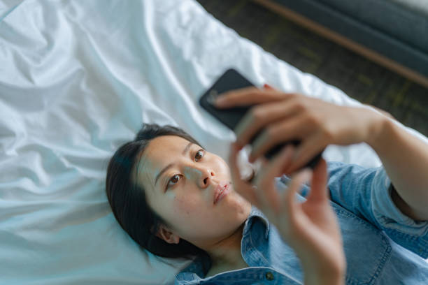 vista de ángulo alto de una mujer joven que usa un teléfono inteligente mientras está acostada en su cama - women solitude enjoyment 20s fotografías e imágenes de stock