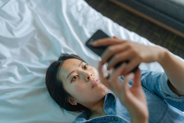 vista ad alto angolo della giovane donna che usa lo smartphone mentre è sdraiata sul suo letto - guilt female women one woman only foto e immagini stock