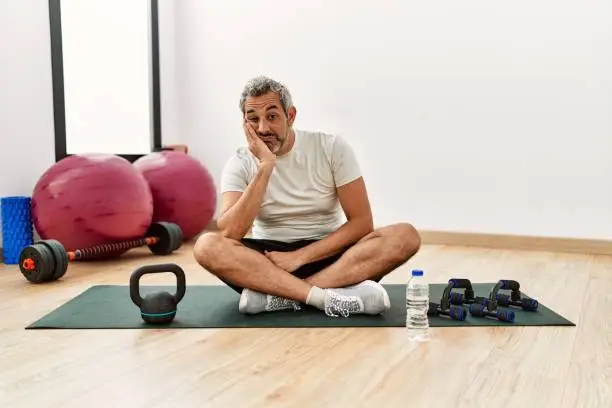 Middle age hispanic man sitting on training mat at the gym thinking looking tired and bored with depression problems with crossed arms.