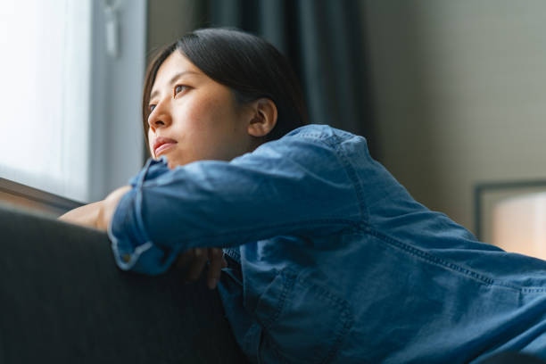 窓越しにソファに座り、家の窓から外を見る若い女性 - 涙が出る 日本人 ストックフォトと画像
