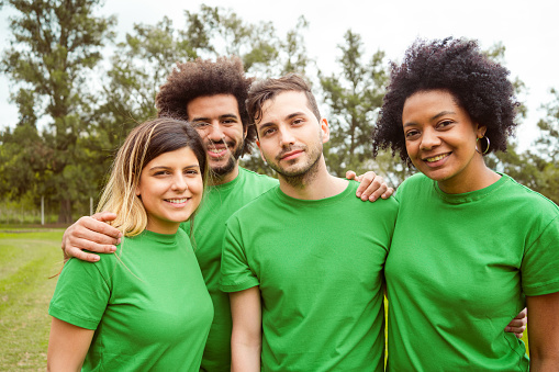 Portrait of confident volunteers standing with arms around. Multiracial group of environmentalists are in park. They are wearing green t-shirts.