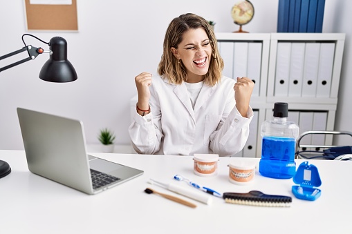 Young hispanic woman working at dentist clinic very happy and excited doing winner gesture with arms raised, smiling and screaming for success. celebration concept.