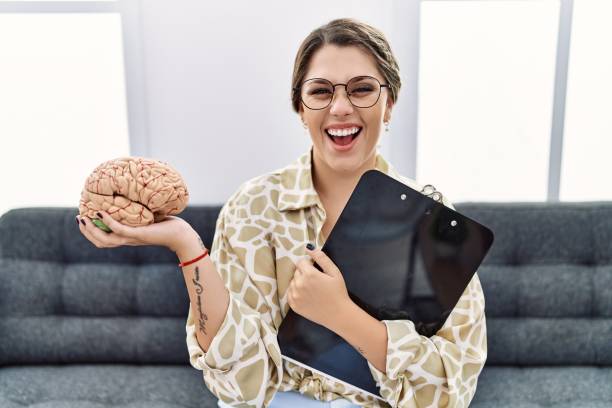 Young hispanic woman holding brain working at psychology clinic smiling and laughing hard out loud because funny crazy joke. Young hispanic woman holding brain working at psychology clinic smiling and laughing hard out loud because funny crazy joke. people laughing hard stock pictures, royalty-free photos & images
