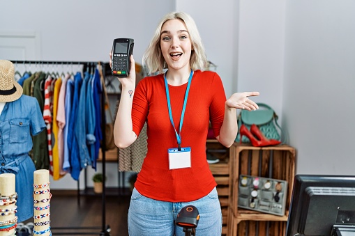 Young caucasian woman working as manager at retail boutique holding dataphone celebrating achievement with happy smile and winner expression with raised hand