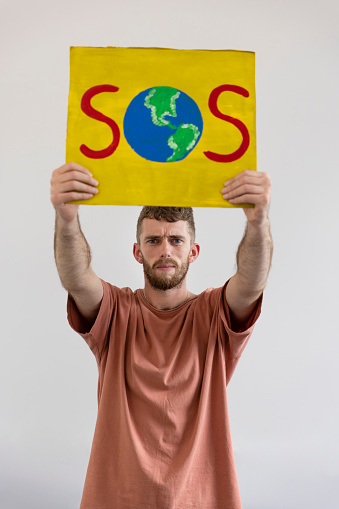 A young man standing in front of a white background holding a handmade sign above his head with 'SOS' on it with a world as the 'O.' The man is looking at the camera with a serious look on his face while holding the sign. He is an activist protesting for climate change.