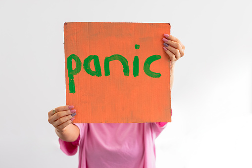An unrecognisable woman standing in front of a white background holding a handmade sign with 'panic' on it in front of her face. She is an activist protesting for climate change.