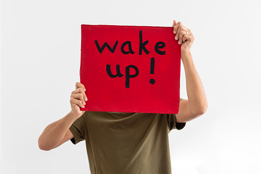 An unrecognisable man holding a handmade sign with 'wake up!' on it up in front of his face while standing in front of a white background. He is an activist protesting for climate change.