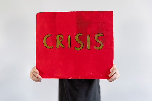 An unrecognisable man standing in front of a white background holding a handmade sign with 'CRISIS' on it in front of his face. He is an activist protesting for climate change.