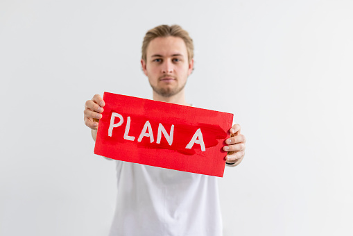 A man standing in front of a white background holding a handmade sign with 'PLAN A' on it. He is looking at the camera with a serious look on his face while holding the sign, the sign is in focus while he isn't. He is an activist protesting for climate change.