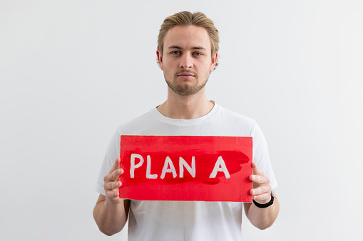A man standing in front of a white background holding a handmade sign with 'PLAN A' on it. He is looking at the camera with a serious look on his face while holding the sign. He is an activist protesting for climate change.
