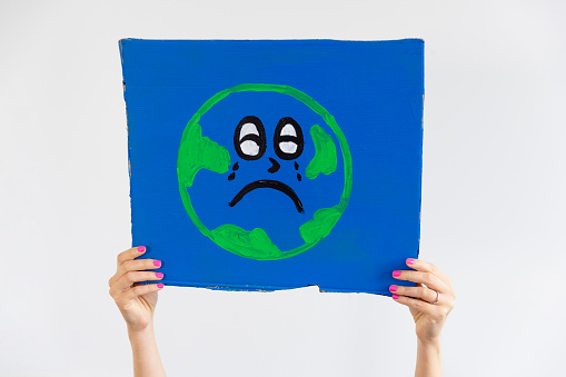 An unrecognisable woman holding a handmade sign with Earth painted on it up in the air. She is an activist protesting for climate change and the Earth has a sad face.