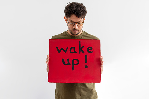 A mid adult man holding a handmade sign with 'wake up!' on it in front of him while standing in front of a white background. He is looking down at the message and is an activist protesting for climate change.