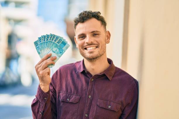 jovem hispânico sorrindo feliz segurando notas reais na cidade. - dinheiro real - fotografias e filmes do acervo