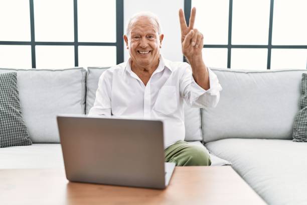 istock Senior man using laptop at home sitting on the sofa smiling looking to the camera showing fingers doing victory sign. number two. 1346576219